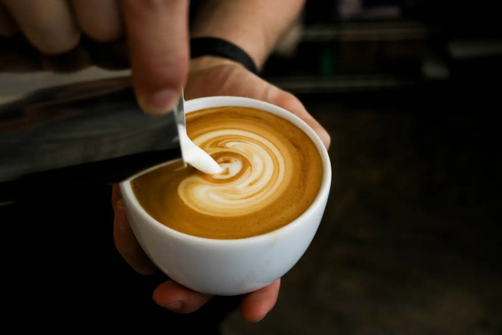 Steamed milk being poured from a metal pitcher into a white cup, forming a leaf pattern
